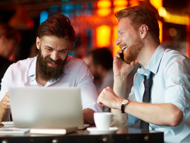 two businessmen talking over coffee, one on a laptop and one talking on the phone